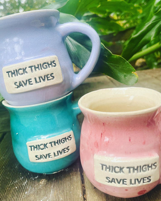 A ceramic coffee mug featuring the empowering phrase ‘Thick Thighs Save Lives,’ celebrating body positivity and self-acceptance.