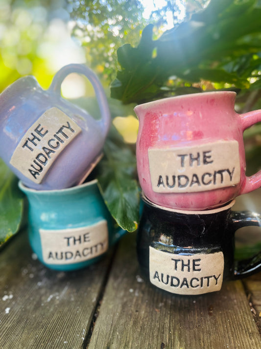 A ceramic coffee mug featuring the empowering phrase ‘The Audacity,’ symbolizing fearless determination and the pursuit of gender equality