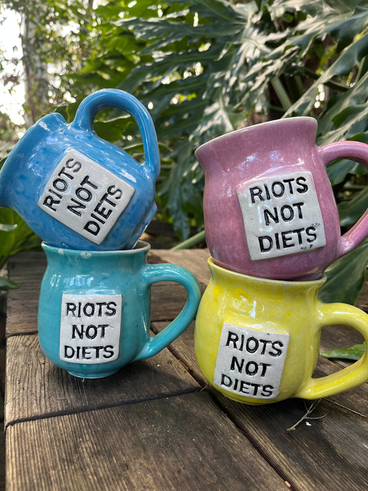 A ceramic coffee mug featuring the empowering phrase ‘Riots Not Diets,’ celebrating body diversity and promoting self-acceptance